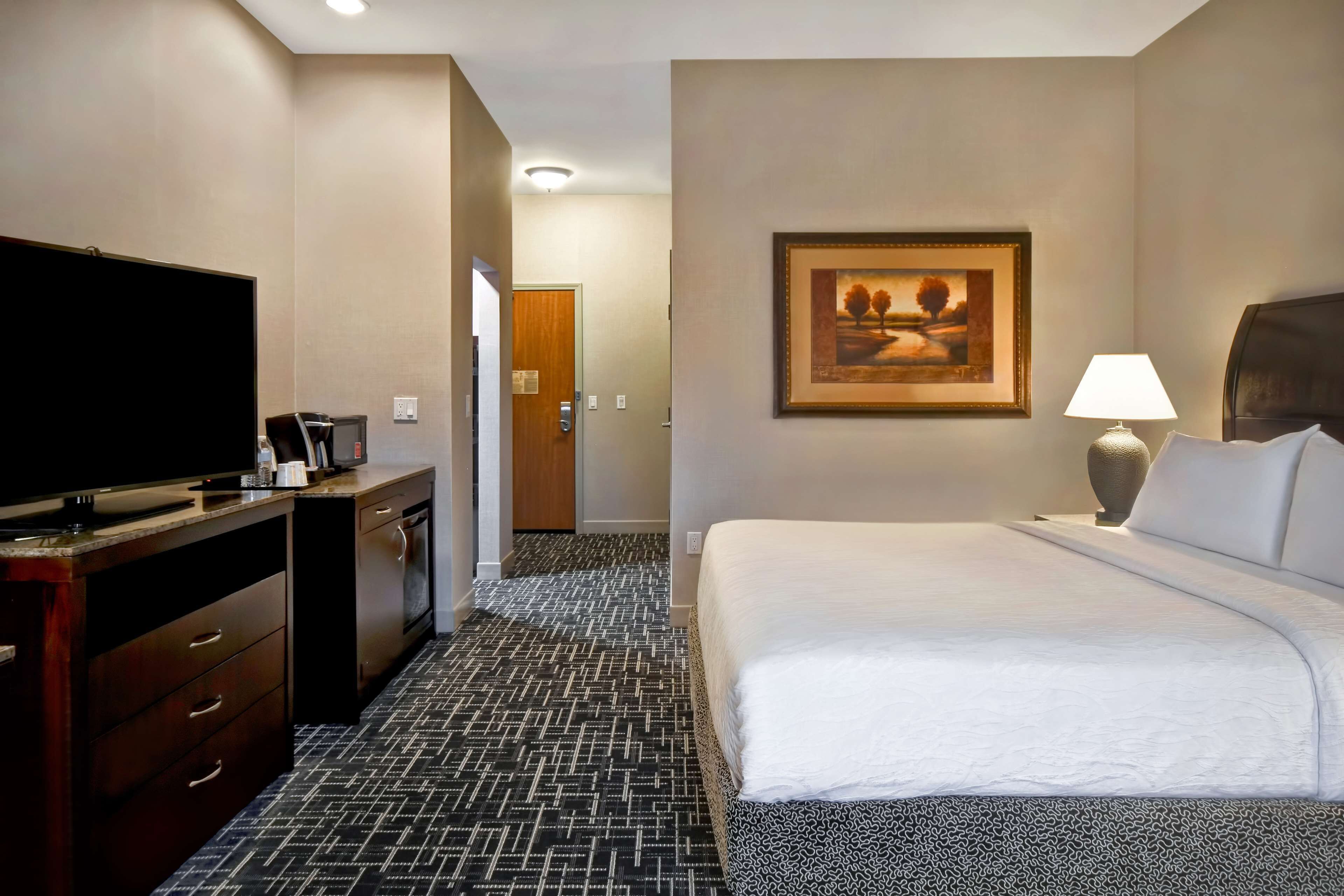 A hotel room with a bed, a TV, a desk with a coffee maker, a lamp, and a framed picture on the wall.