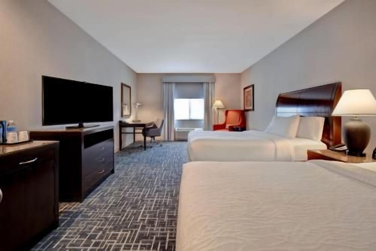 A hotel room with two beds, a desk, a television, and a window.