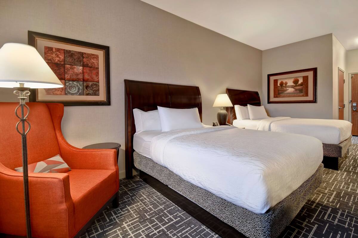 A hotel room with a large bed, a smaller bed, an orange armchair, and two bedside tables with lamps.