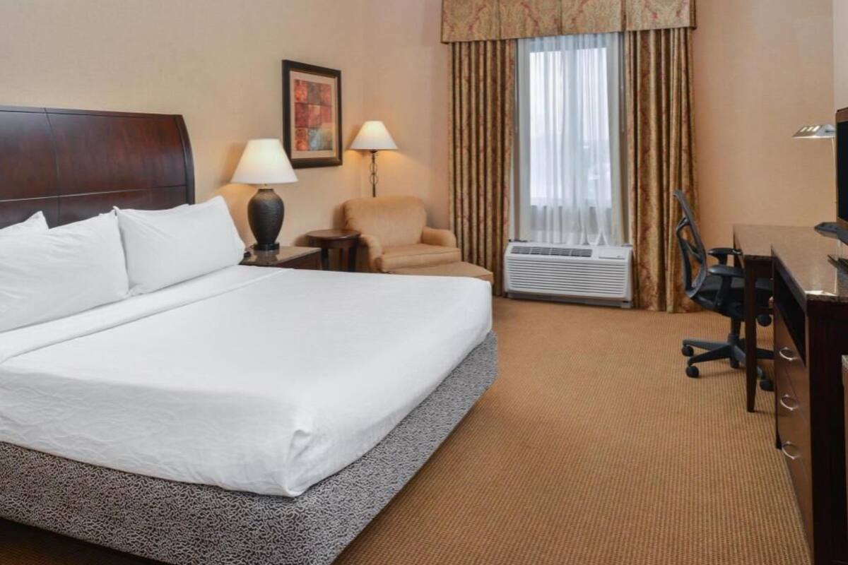 A hotel room with a large bed, a desk, a chair, a lamp, an air conditioner, and a window with curtains.