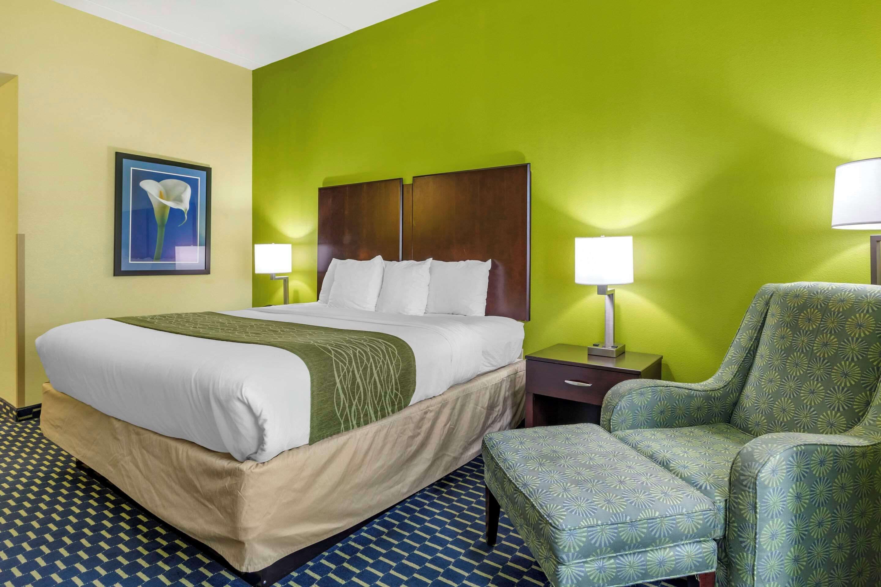 Quarto de hotel com uma cama grande, duas luminárias de cabeceira, uma poltrona verde e uma moldura na parede.