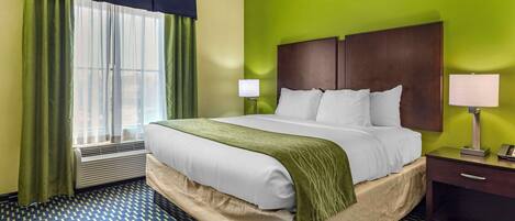 A hotel room with a large bed, green walls, a window with curtains, a bedside table with a lamp, and a patterned floor.