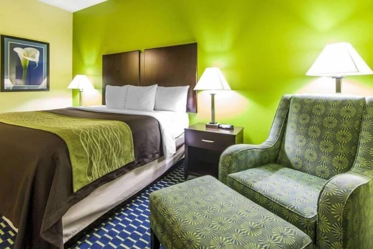 A hotel room with a bed, two bedside tables with lamps, a green patterned armchair, and a patterned ottoman.