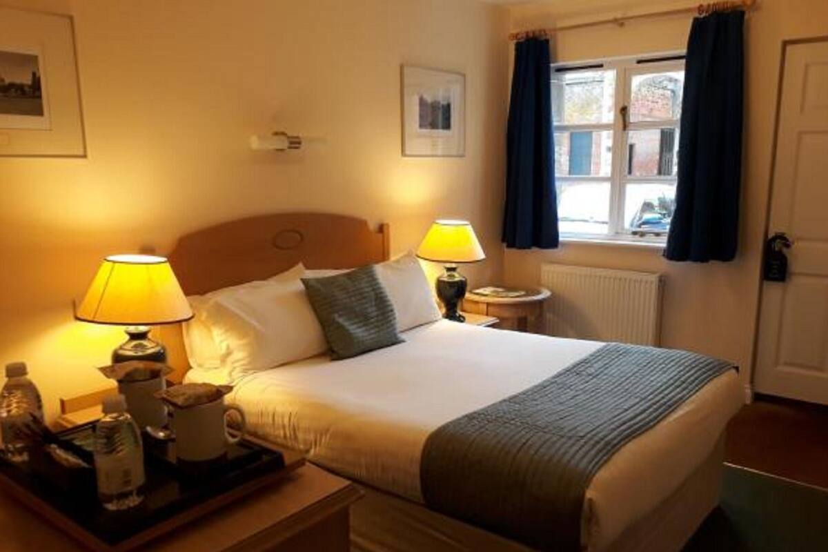 Hotellihuone, jossa on sänky, kaksi yöpöytävalaisinta, yöpöytä, jossa on tarjoitinalusta hygieniatuotteille, ja ikkuna, jossa on verhot.