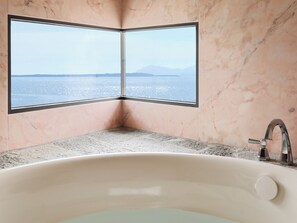 A bathroom with a bathtub, a sink, and a large window offering a view of the sea and mountains.