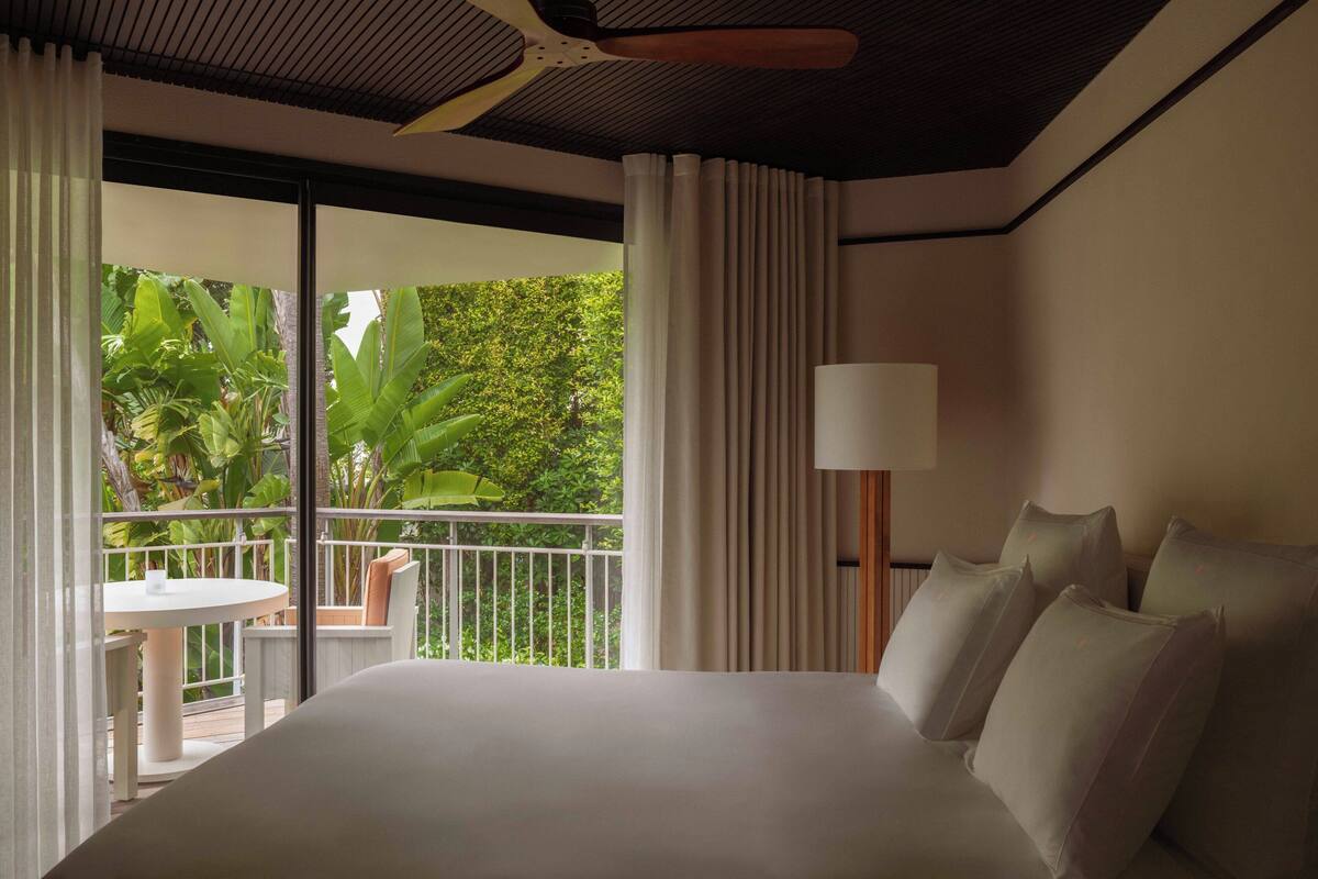 A hotel room with a large bed, a balcony with a table and chairs, and a view of lush greenery.