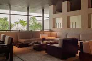 A modern living room with a sectional sofa, a coffee table, and a lamp. There are large windows with a view of greenery outside.