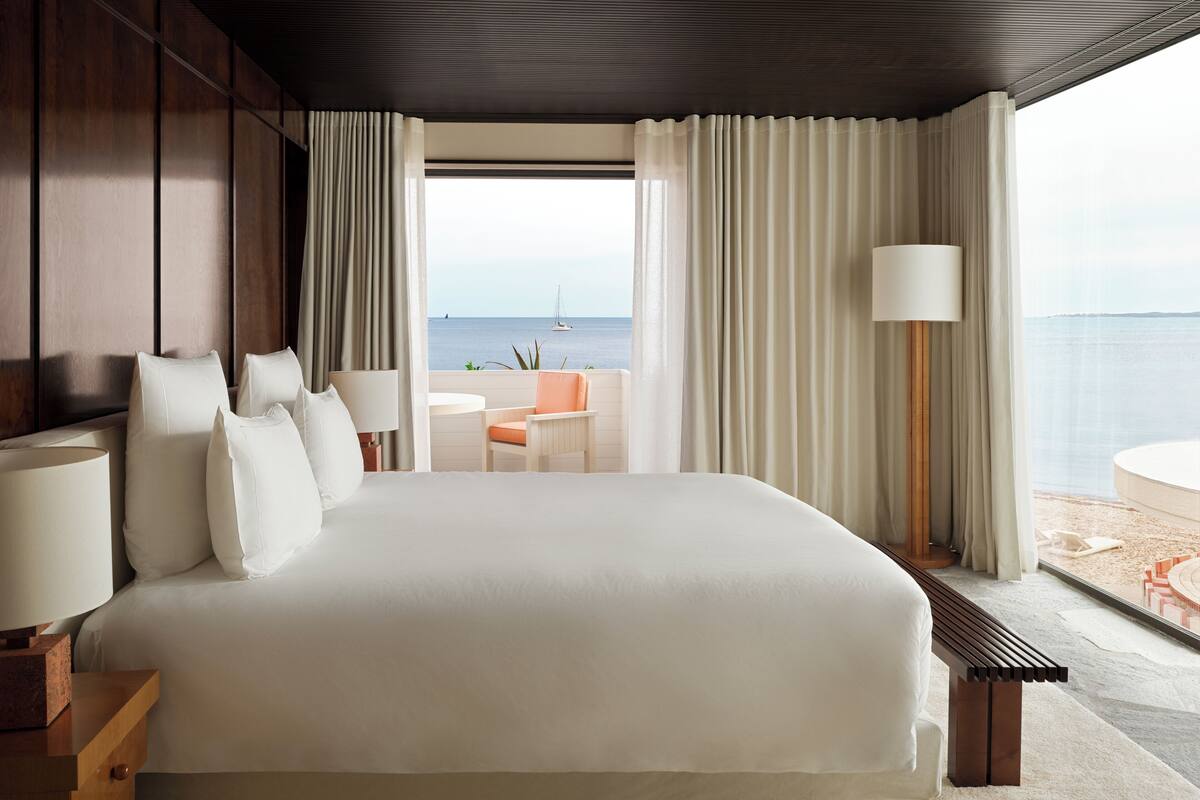 A hotel room with a large bed, a seating area with a view of the sea, and a balcony with a table and chairs.