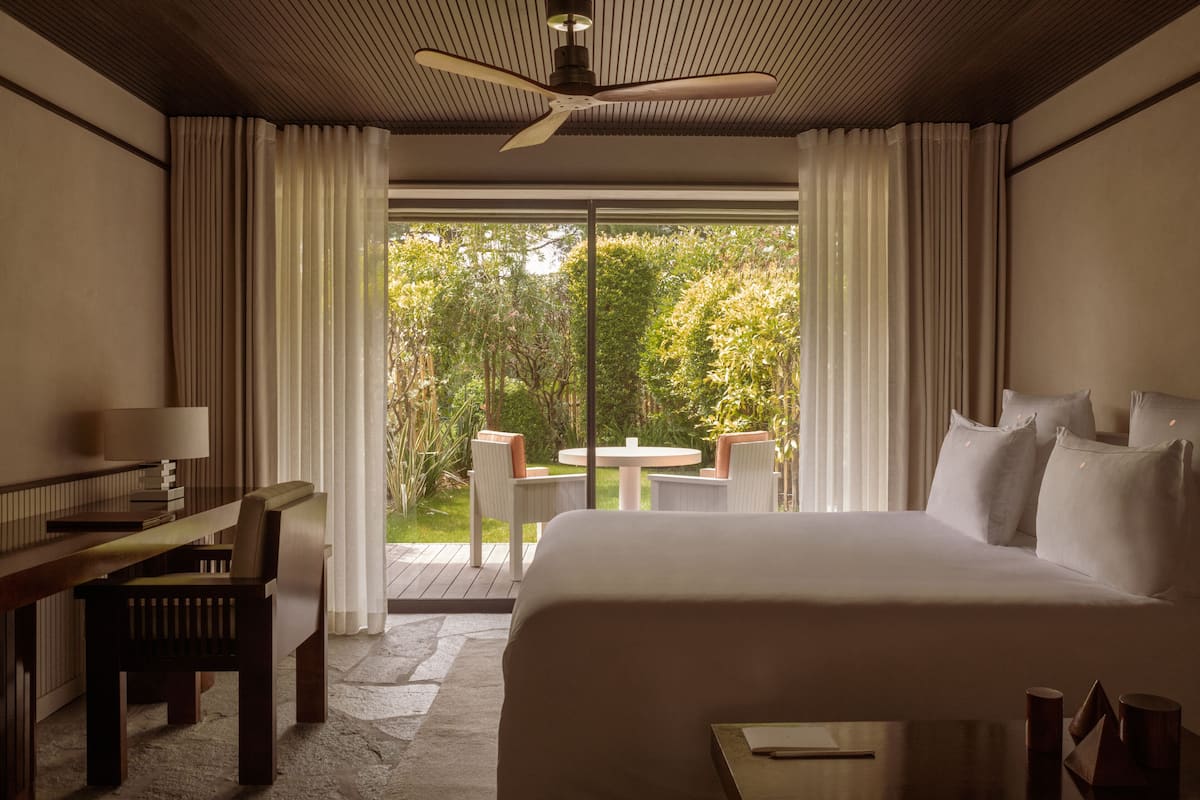 A hotel room with a large bed, a desk with a chair, a small table, and a balcony with a view of greenery.
