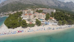 Vista aérea de un hotel costero con varias construcciones, una playa y montañas al fondo.