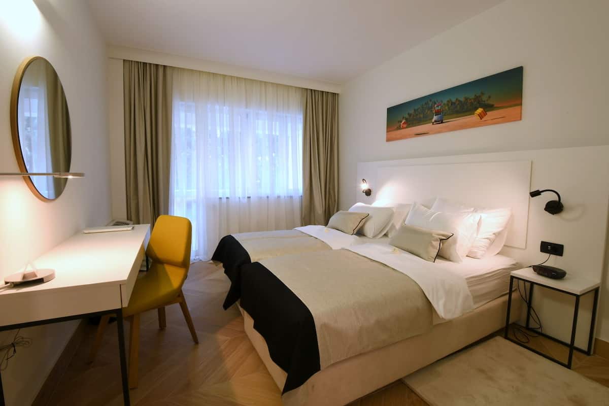 A hotel room with a bed, a desk, a chair, a painting, and a window with curtains.