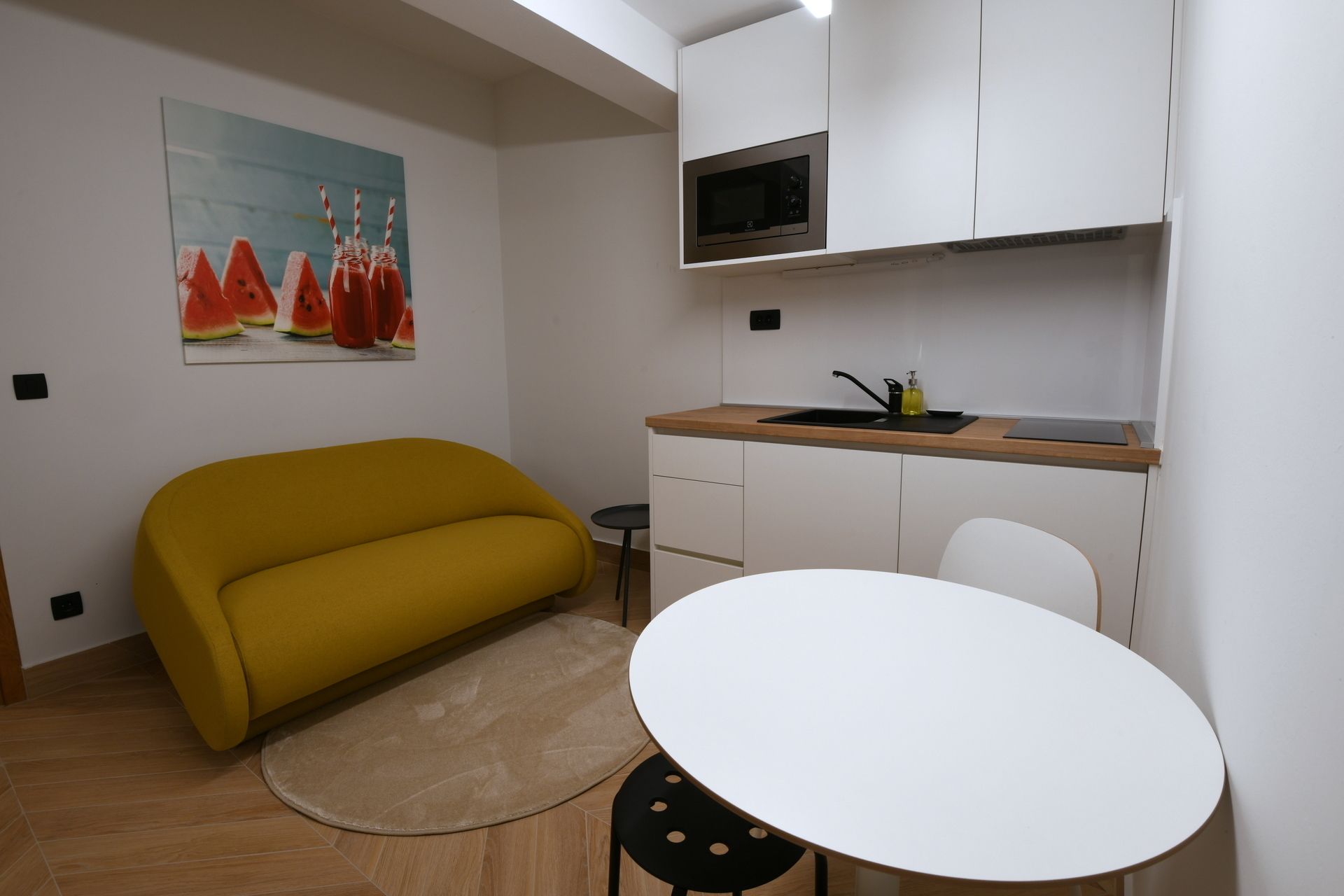 A compact living space with a yellow sofa, a white table, a microwave, and a small kitchen area.