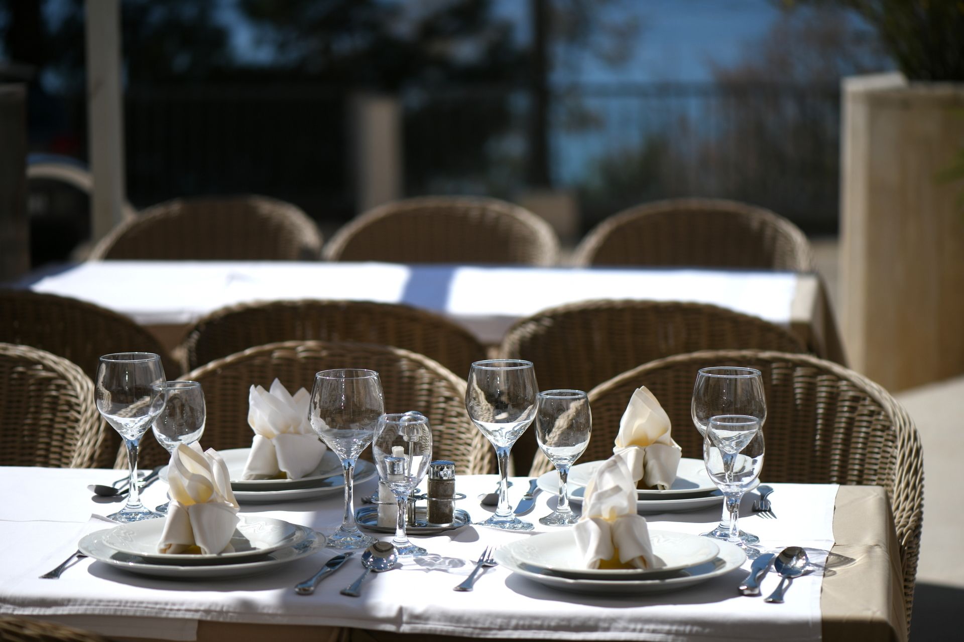 A dining table set with white plates, clear wine glasses, and silverware, surrounded by wicker chairs.