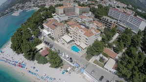 Vista aérea de un hotel costero con varias edificaciones, una piscina y una playa con sombrillas.