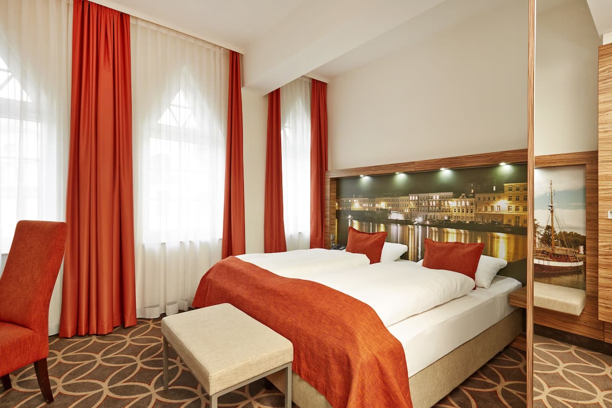 Ein Hotelzimmer mit einem großen Bett, zwei roten Sesseln, einem kleinen Tisch und Blick auf die Stadt.