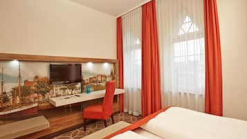 Habitació d'hotel amb un llit, una taula de treball amb cadira, televisió i finestra amb cortines.