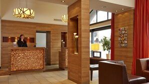 A modern reception area with a wooden reception desk, seating area, and large windows. - H+ Hotel Lübeck (Lübeck)