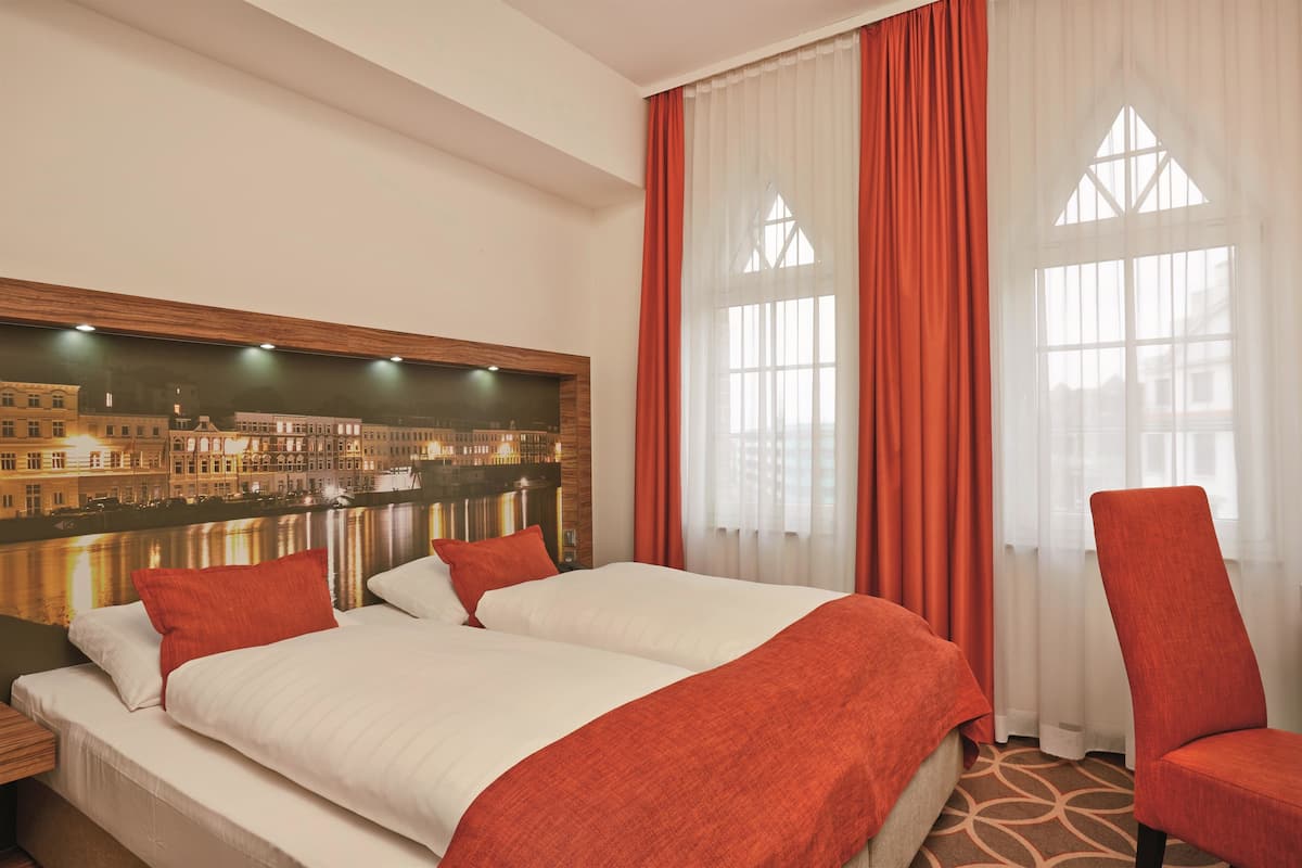 Ein Hotelzimmer mit einem großen Bett, zwei Kissen, einem roten Sessel, einem Nachttisch und einem an der Wand befestigten Fernseher, der eine Stadtansicht zeigt.