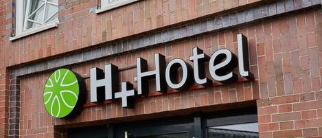 A hotel sign with a green logo and the name "H+Hotel" on a brick building.