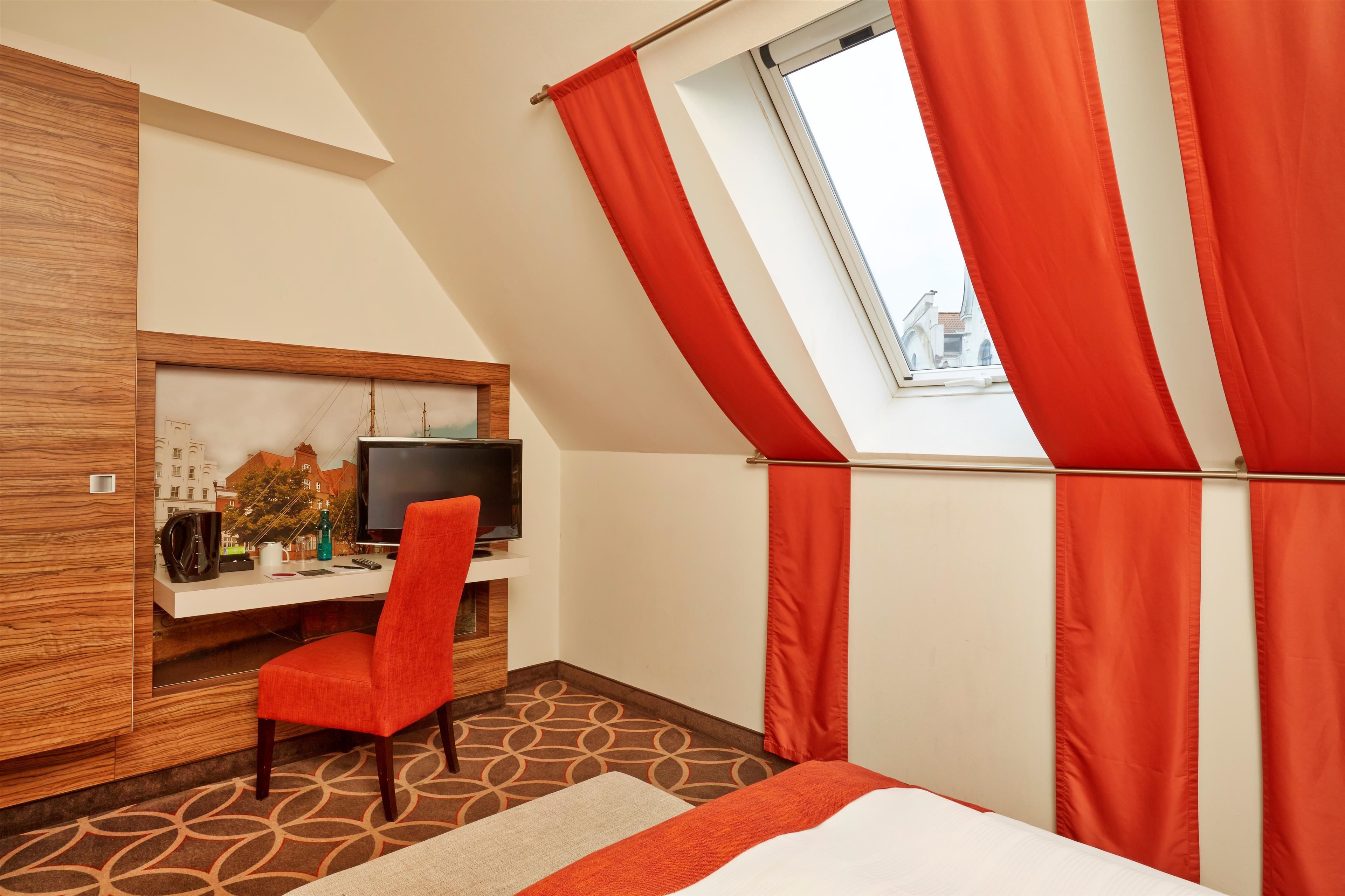 A hotel room with a bed, a desk with a TV, a chair, and a window with red curtains.