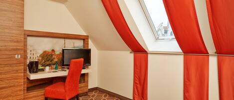 Habitación de hotel con cama, escritorio con televisor, una silla y ventana con cortinas rojas.