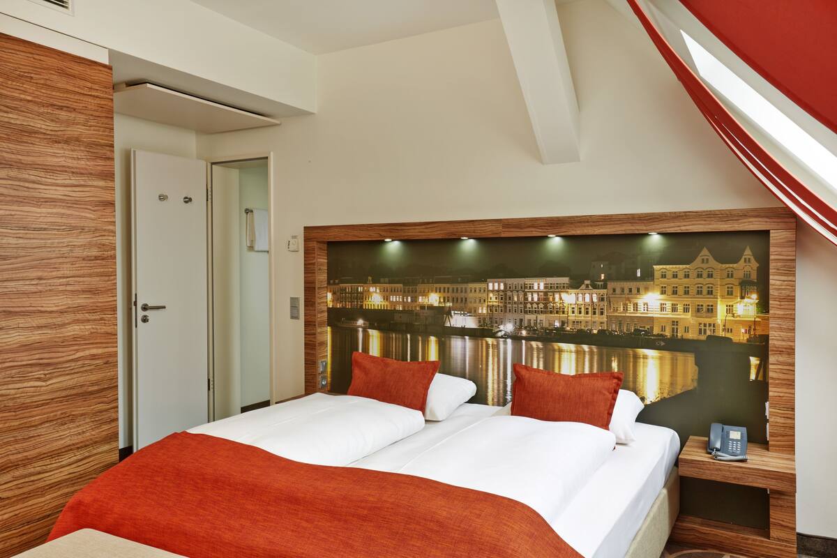 Ein Hotelzimmer mit einem großen Bett, einem hölzernen Kopfteil, einem Nachttisch mit Telefon und Blick auf eine Stadtlandschaft durch das Fenster.