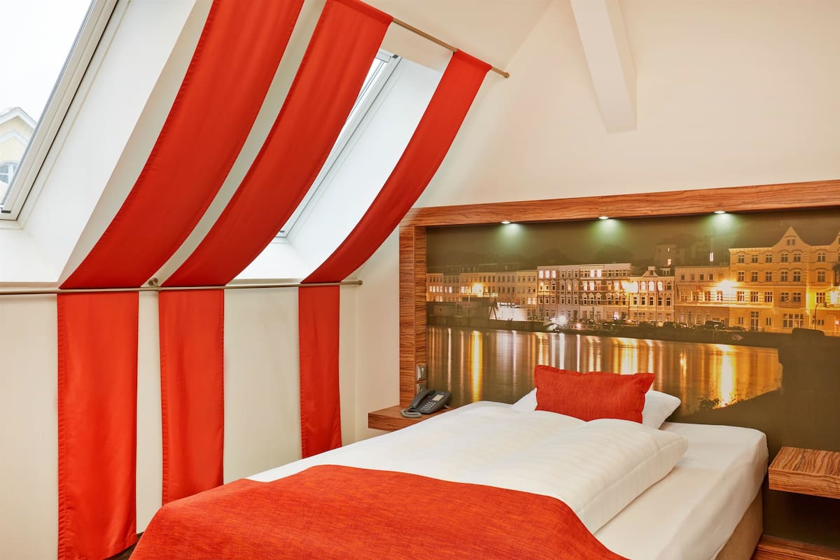 Ein Hotelzimmer mit einem Bett, einem Nachttisch mit Telefon, einem Fenster mit roten Vorhängen und einem Stadtbild-Wandgemälde.