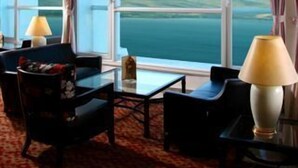 Une chambre d’hôtel dotée d’une grande fenêtre offrant une vue panoramique sur la mer et les collines. On y trouve deux fauteuils et une petite table avec une lampe.