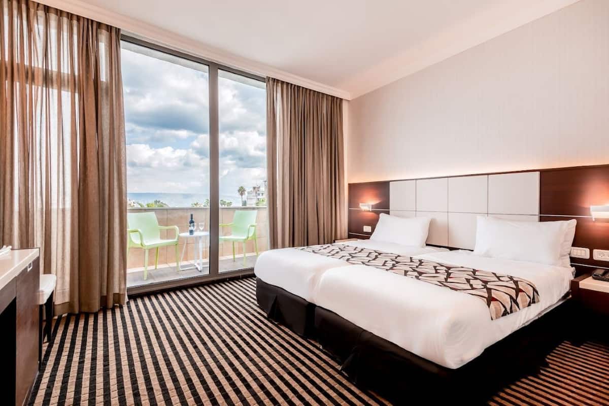 Habitación de hotel con una cama grande, un escritorio, una silla y un balcón con vistas a la ciudad.