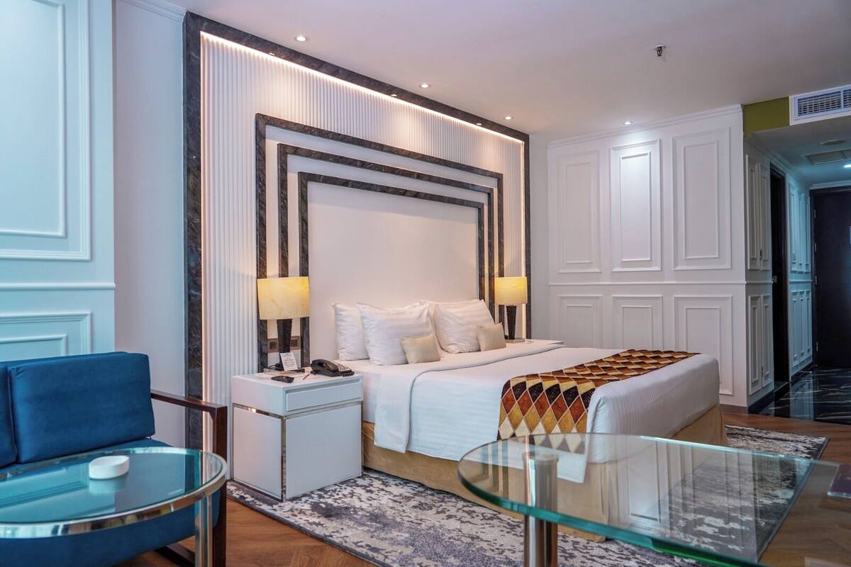 A modern hotel room with a large bed, bedside lamps, a glass table, and a blue chair.