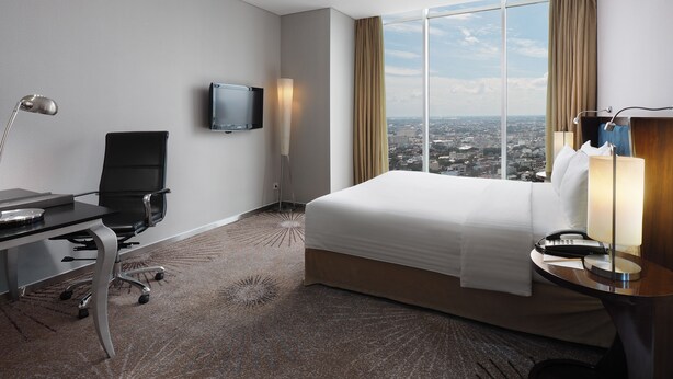 A hotel room with a bed, desk, chair, TV, and a view of the city. - Cambridge Hotel Medan (Medan)