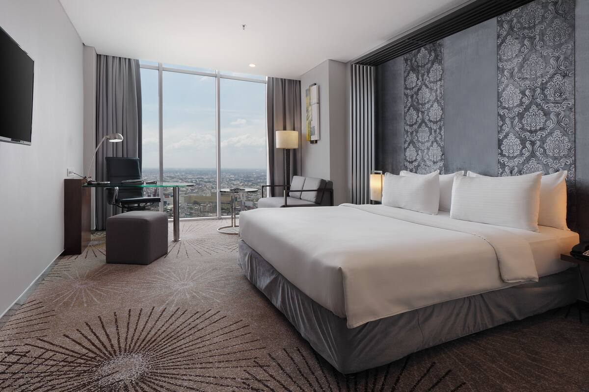 A modern hotel room with a large bed, a desk with a laptop, a chair, and a view of the city through large windows.