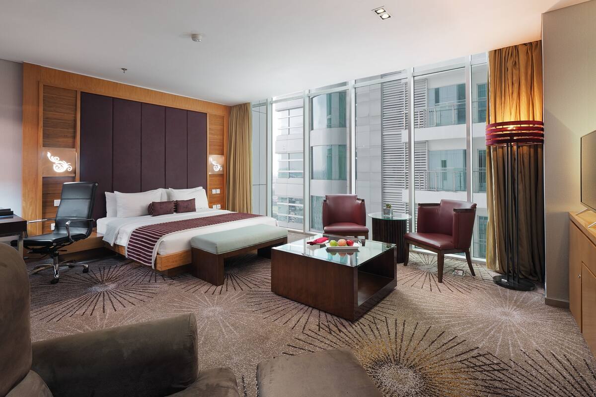 A modern hotel room with a large bed, two armchairs, a coffee table, and a view of the city through large windows.
