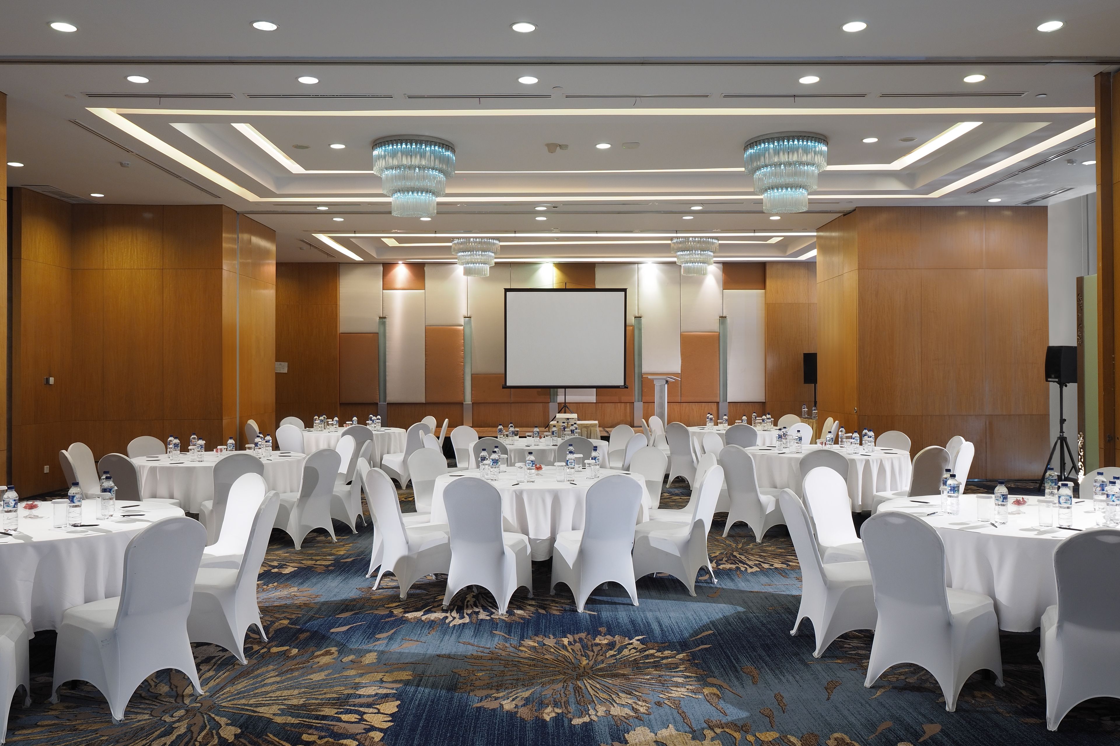 Un salón de conferencias con mesas redondas, sillas blancas y una pantalla grande.