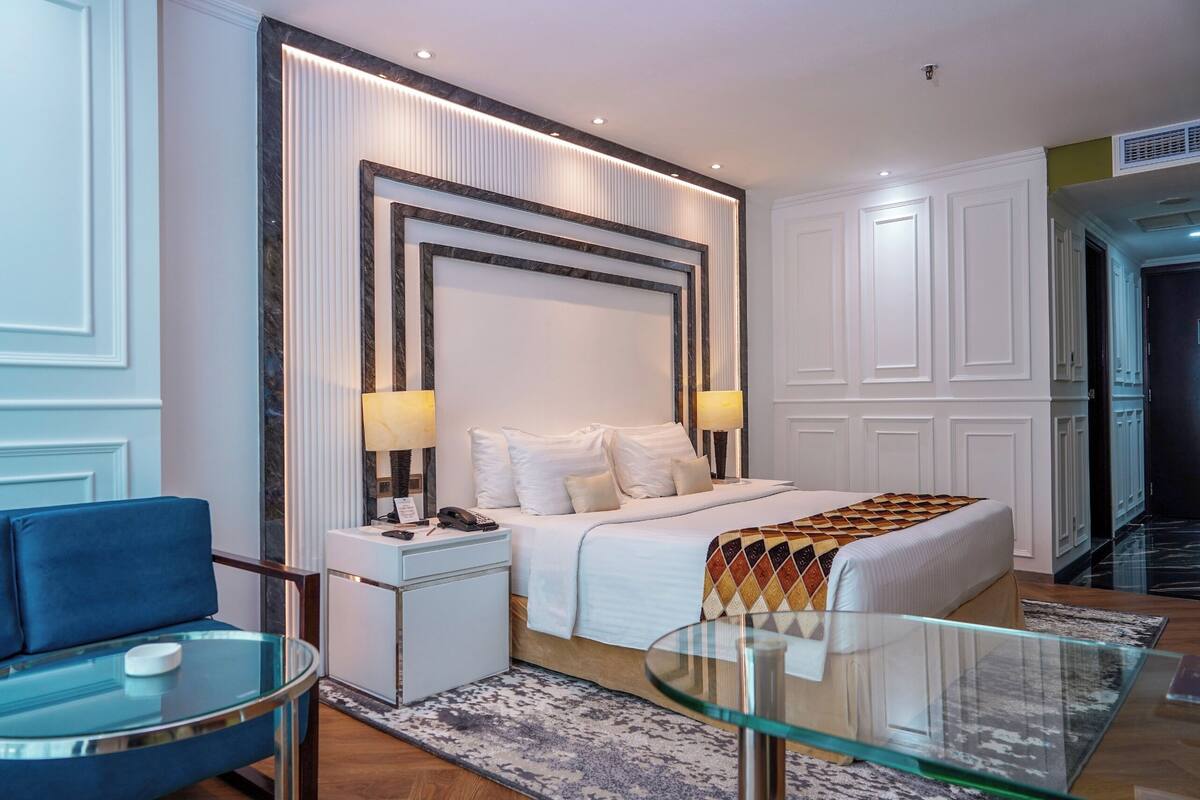 A modern hotel room with a large bed, bedside lamps, a glass table, a blue armchair, and a patterned throw blanket.