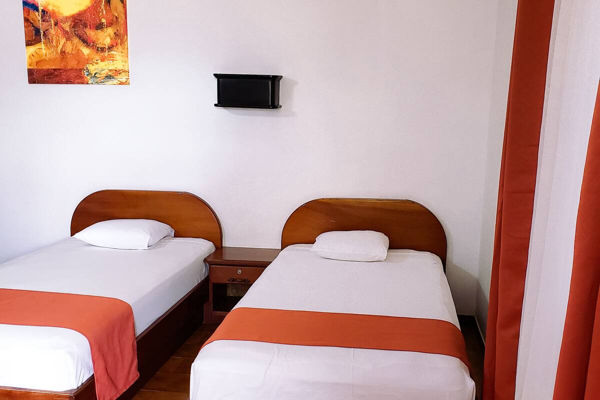 A room with two single beds, each with a wooden headboard and white bedding. There is a small bedside table between the beds, and a wall-mounted TV above it. The room has a tiled floor and a painting on the wall.
