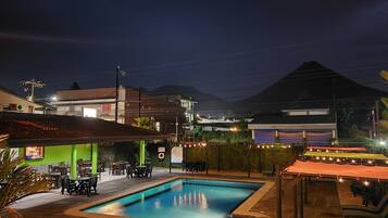Zona de piscina con sillones reclinables y mesas, rodeada de edificios y una montaña al fondo.