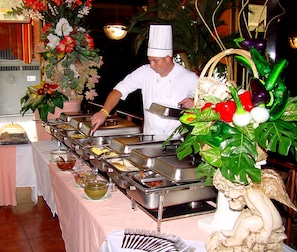 A buffet setup with multiple food stations, a chef serving food, and decorative plants. - Hotel Cervantes (Guadalajara)