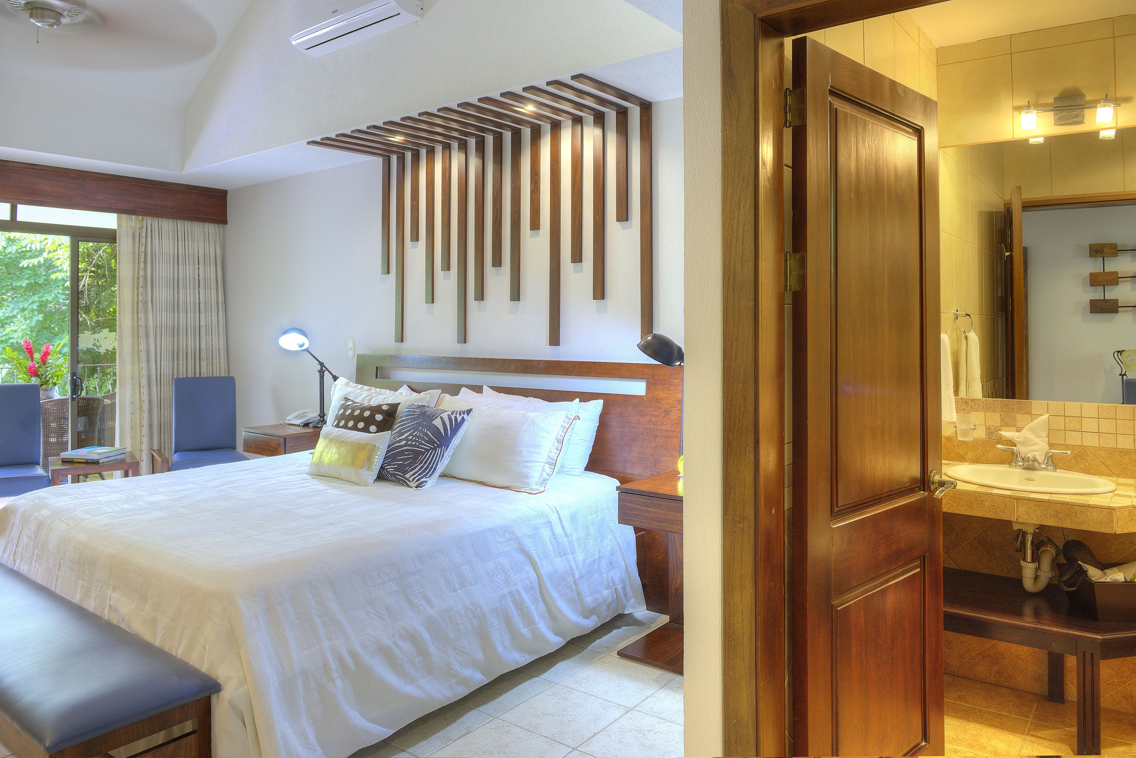 A hotel room with a large bed, a bedside table, a lamp, and a view of the outdoors.