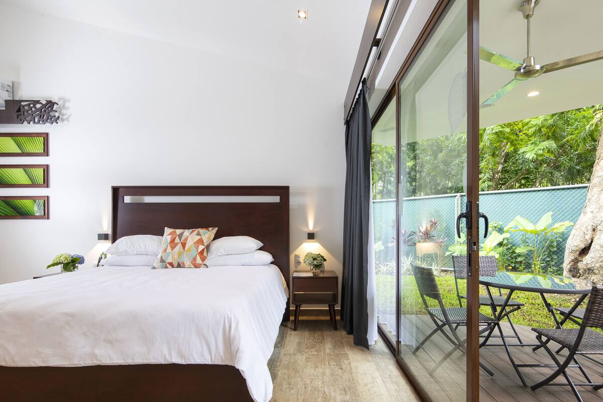 A bedroom with a bed, bedside table, and a view of an outdoor patio with chairs and greenery.