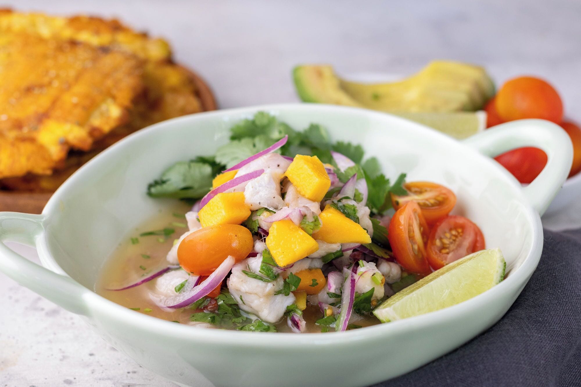 A bowl of ceviche with mango, tomato, and onion, accompanied by a lime wedge and a slice of avocado.