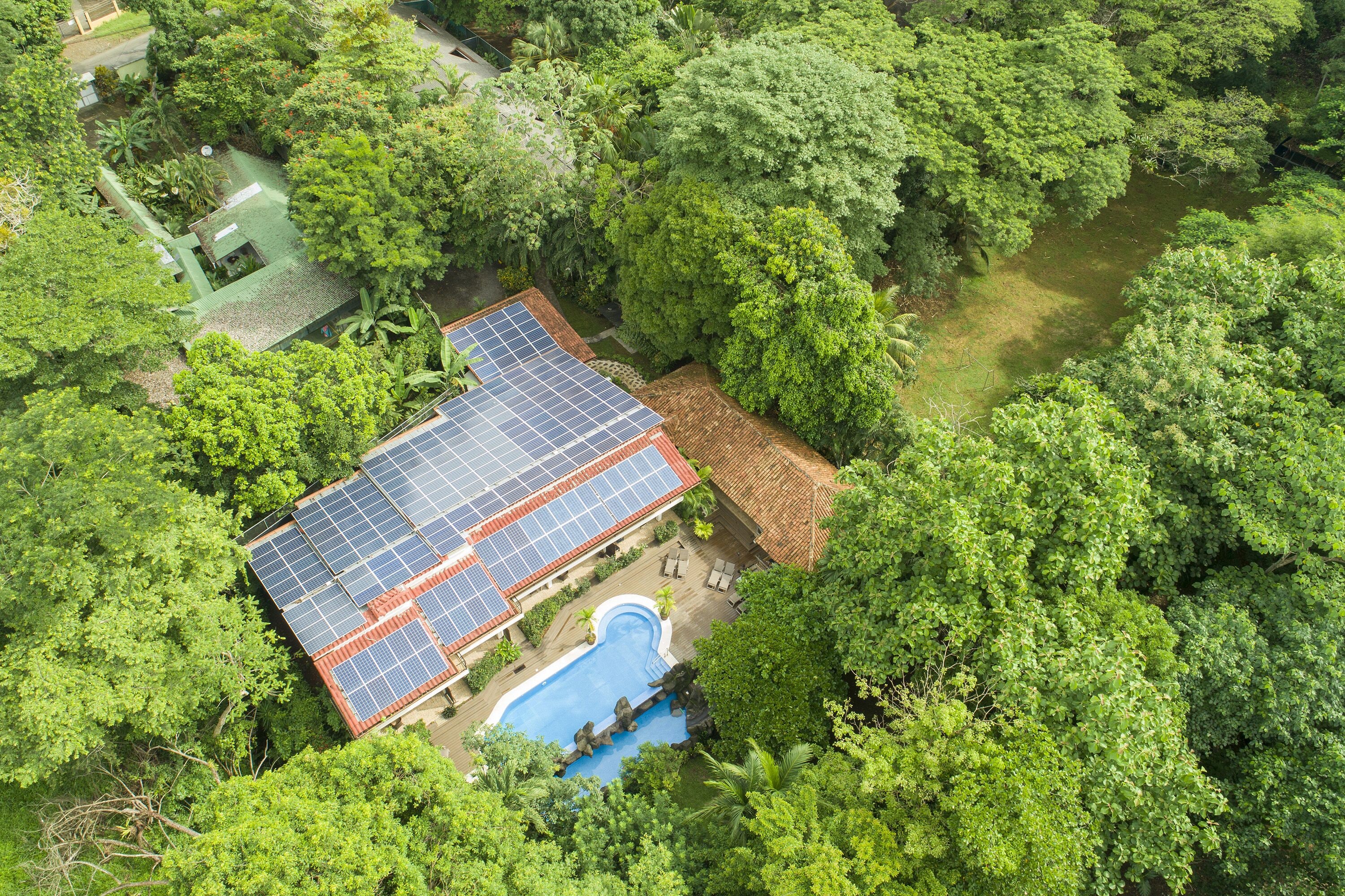 A house with a solar panel roof, surrounded by dense greenery and a swimming pool.