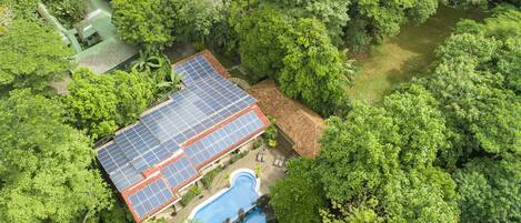 A house with a solar panel roof, surrounded by dense greenery and a swimming pool.