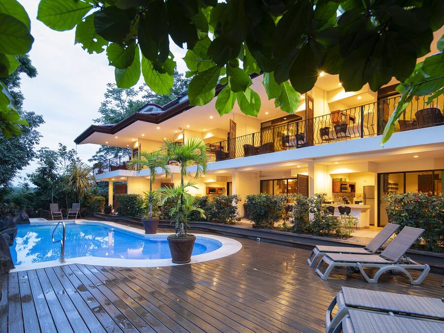 Una casa moderna de dos pisos con piscina, mobiliario de exterior y abundante vegetación.
