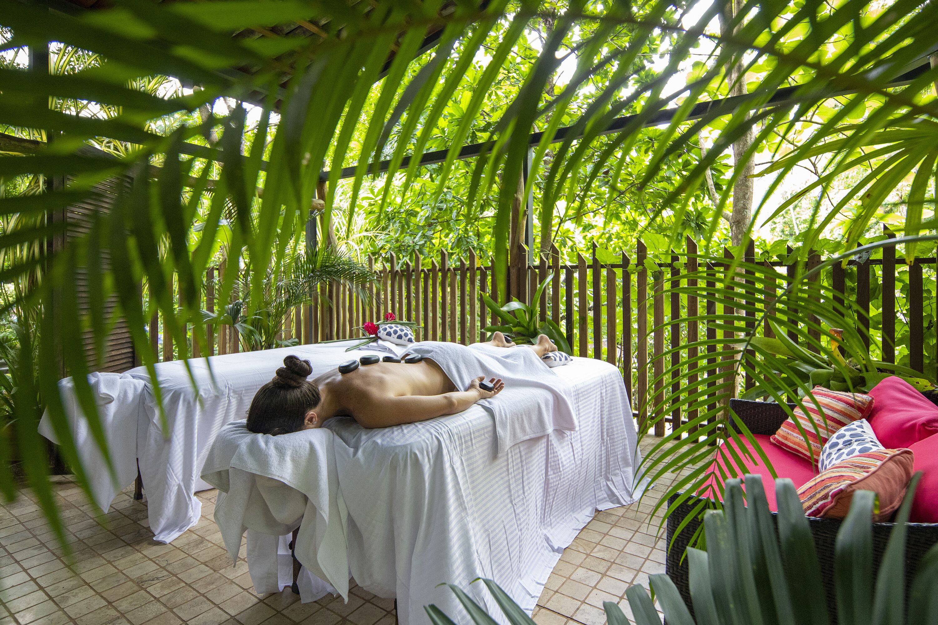 A spa area with two massage tables covered in white linens, surrounded by lush green foliage and a wooden fence.