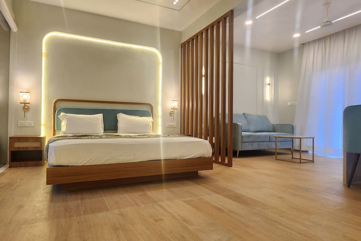 Una habitación de hotel moderna con una cama de madera, una zona de estar con una silla azul y un ventanal con cortinas.