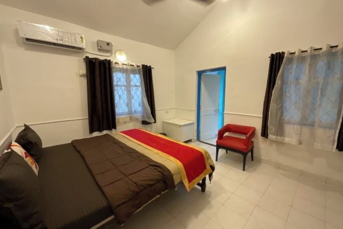 A bedroom with a bed, a chair, an air conditioning unit, a window with curtains, and a door.