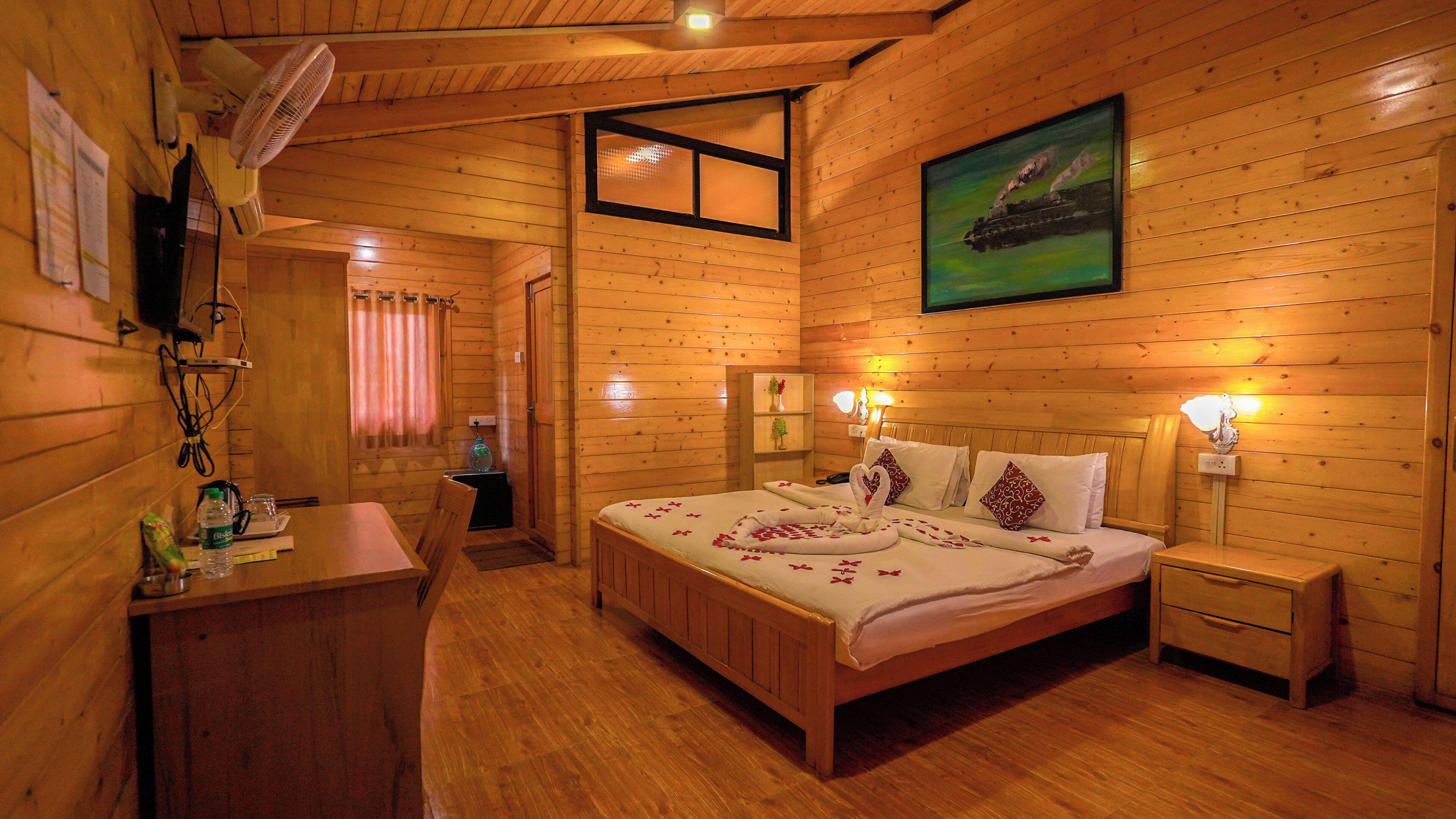 Une chambre dans une cabane en bois, avec un lit, des tables de chevet, un bureau, une chaise, un ventilateur, une fenêtre et un tableau accroché au mur.