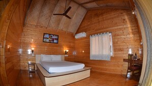 A cozy wooden cabin room with a bed, a ceiling fan, a small table with a chair, and a window with curtains. - Ala Goa Resort (Betalbatim)
