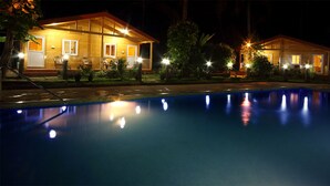 A nighttime view of a hotel with two wooden buildings, a swimming pool, and surrounding greenery. - Ala Goa Resort (Betalbatim)
